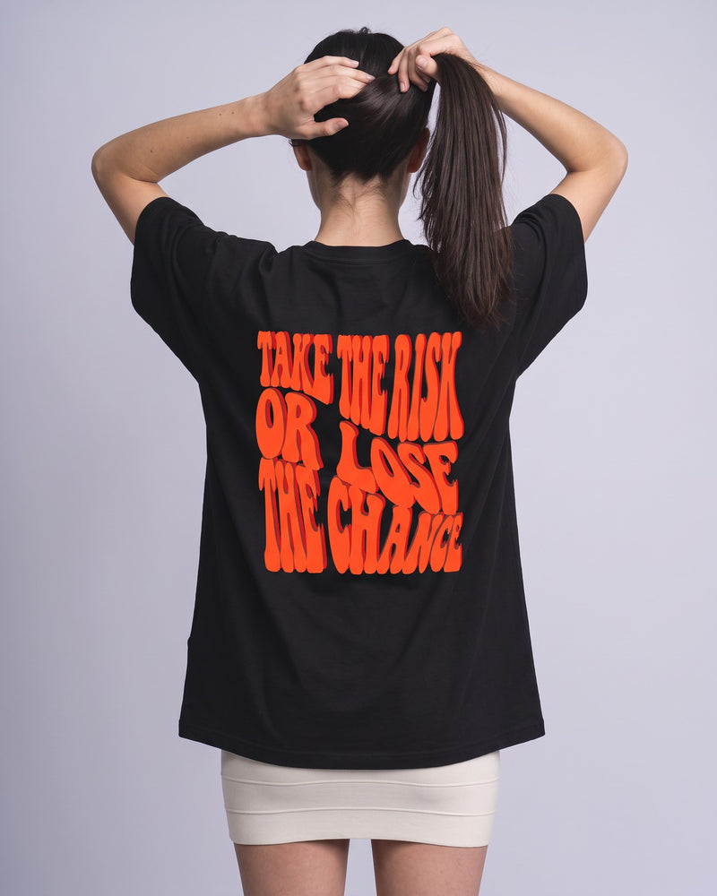 Camiseta oversized feminina MALTA® take the risk preta vista de costas no estúdio, com a frase “take the risk or lose the chance”