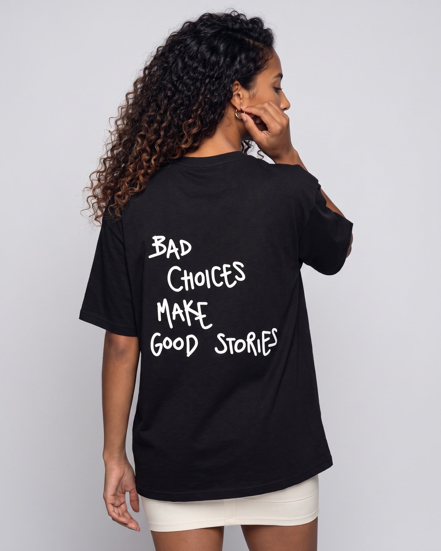 Camiseta oversized feminina MALTA® bad choices preta vista de costas no estúdio, com a frase “bad choices make good stories”