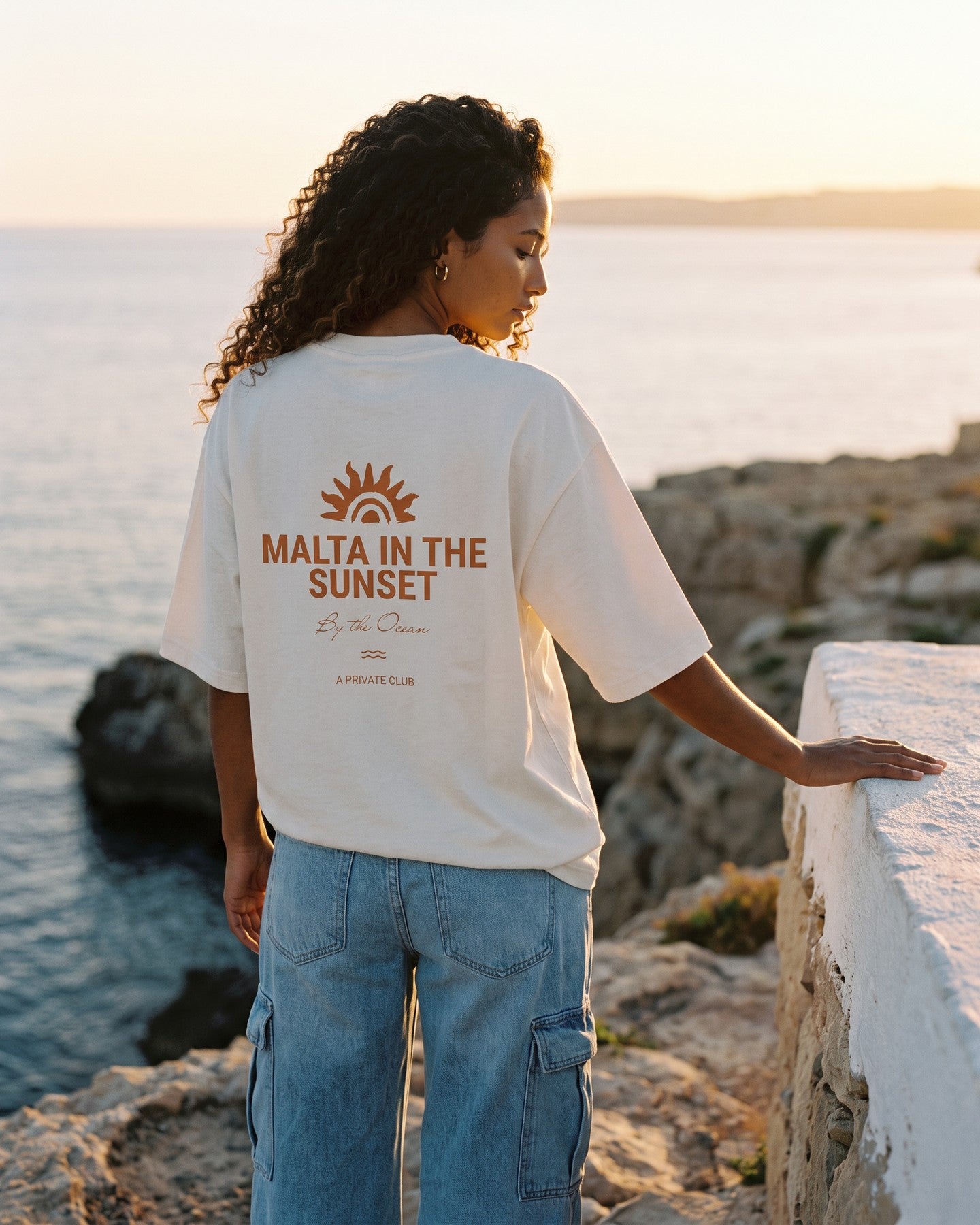 Mulher de costas usando a camiseta oversized MALTA® innerbloom off-white na costa rochosa ao pôr do sol