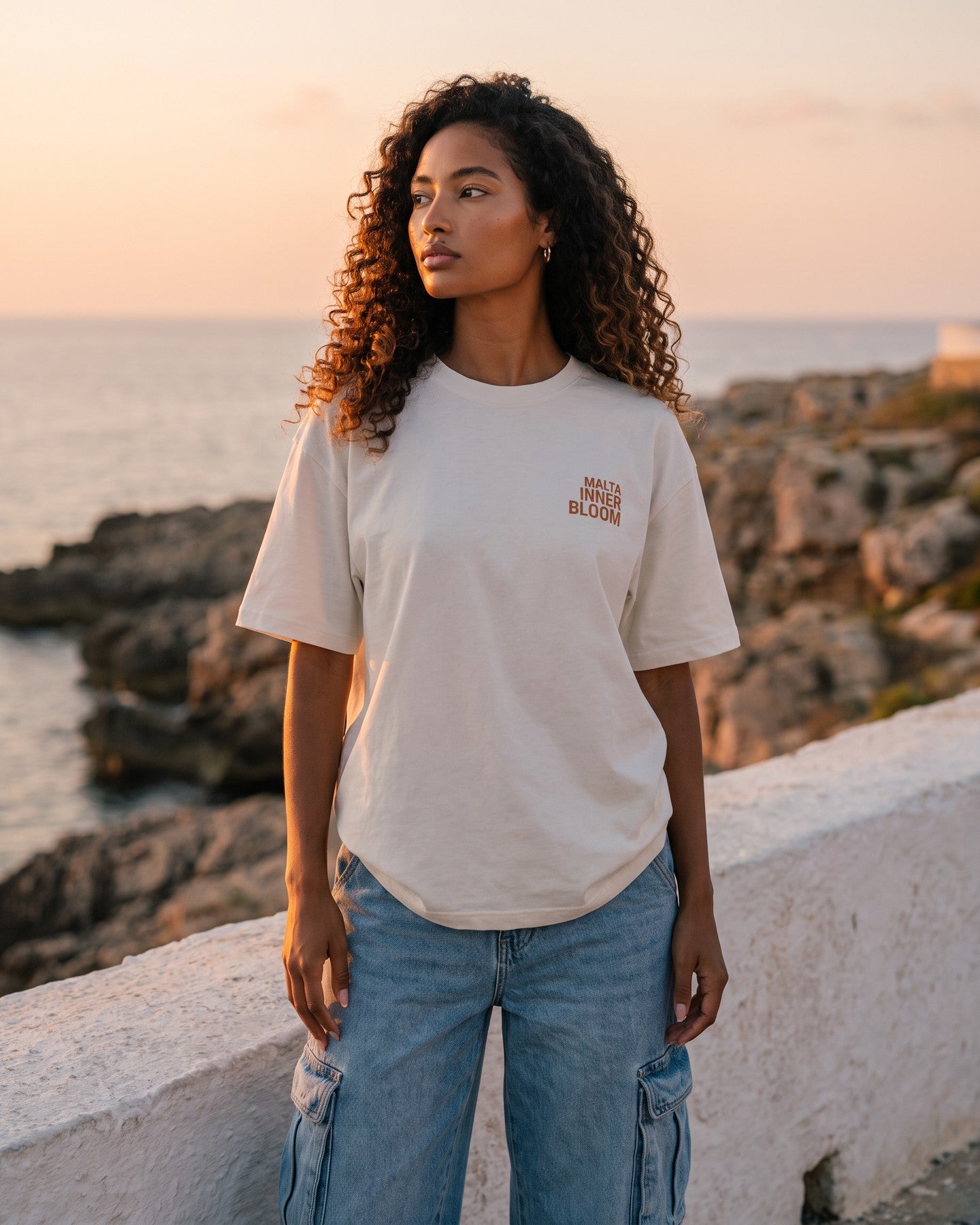 Mulher usando a camiseta oversized MALTA® innerbloom off-white na costa rochosa ao pôr do sol
