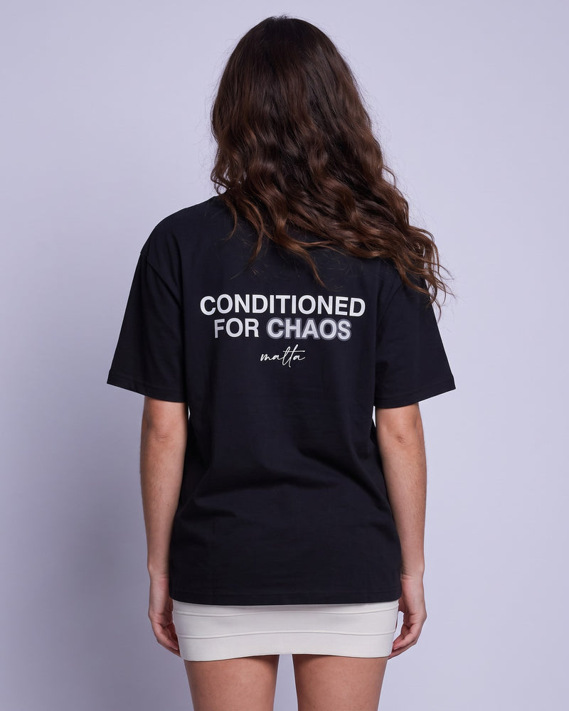 Camiseta oversized feminina MALTA® conditioned for chaos preta vista de costas no estúdio, com a frase “conditioned for chaos”
