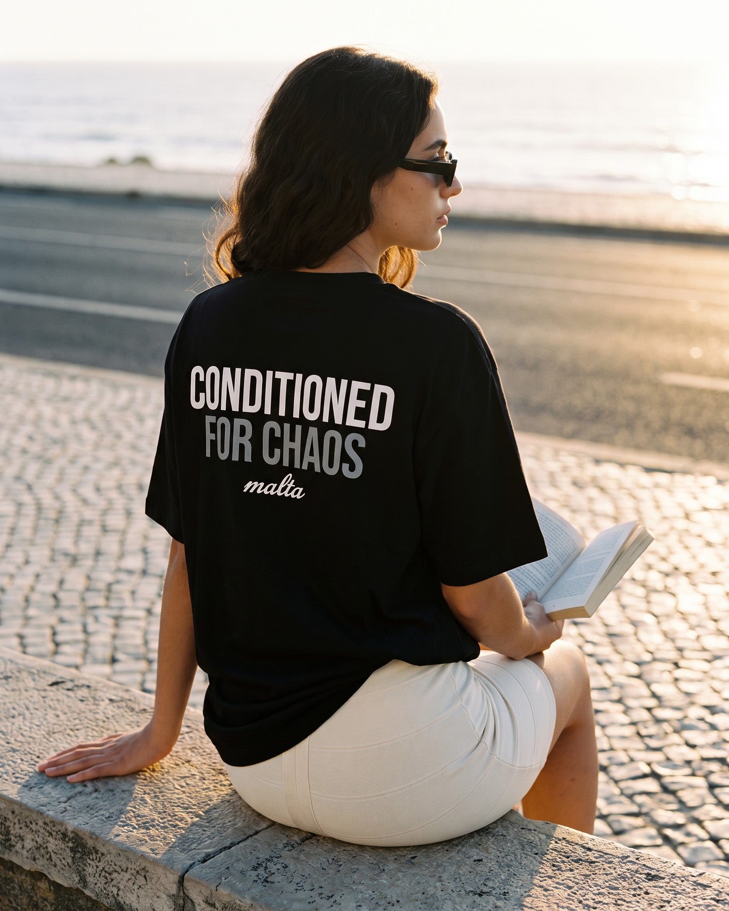 Mulher de costas usando a camiseta oversized MALTA® conditioned for chaos preta num calçadão de pedra portuguesa