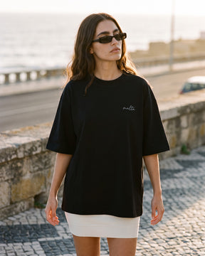 Mulher usando a camiseta oversized MALTA® conditioned for chaos preta num calçadão de pedra portuguesa