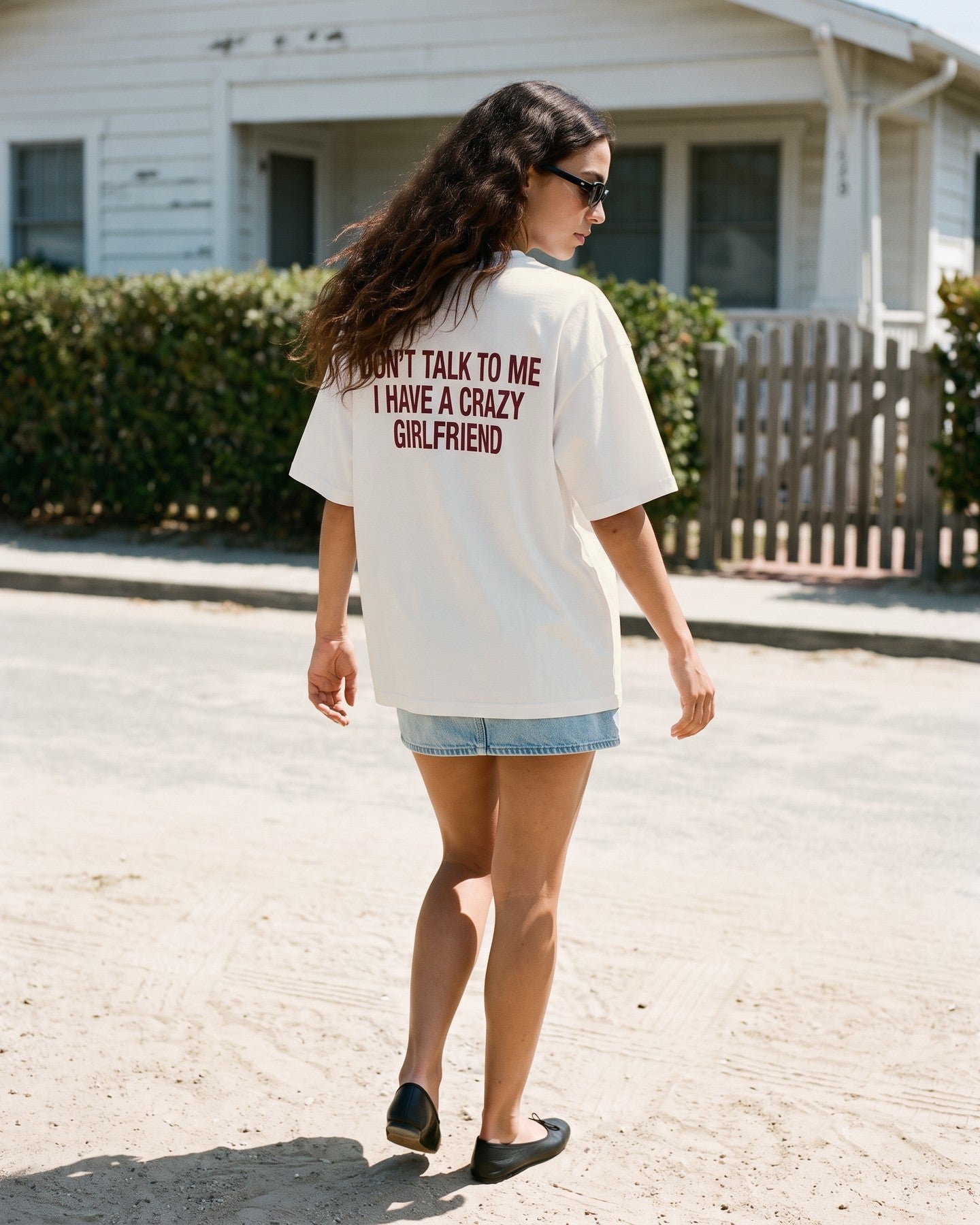 Mulher de costas usando a camiseta oversized MALTA® crazy girlfriend off-white diante de uma casa de praia