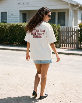 Mulher de costas usando a camiseta oversized MALTA® crazy girlfriend off-white diante de uma casa de praia