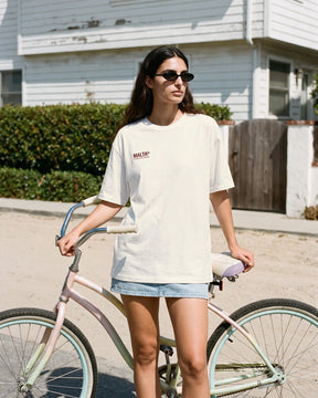 Mulher usando a camiseta oversized MALTA® crazy girlfriend off-white diante de uma casa de praia