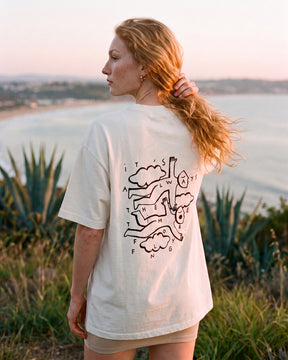 Mulher de costas usando a camiseta oversized MALTA® flying off-white numa encosta sobre a baía ao pôr do sol