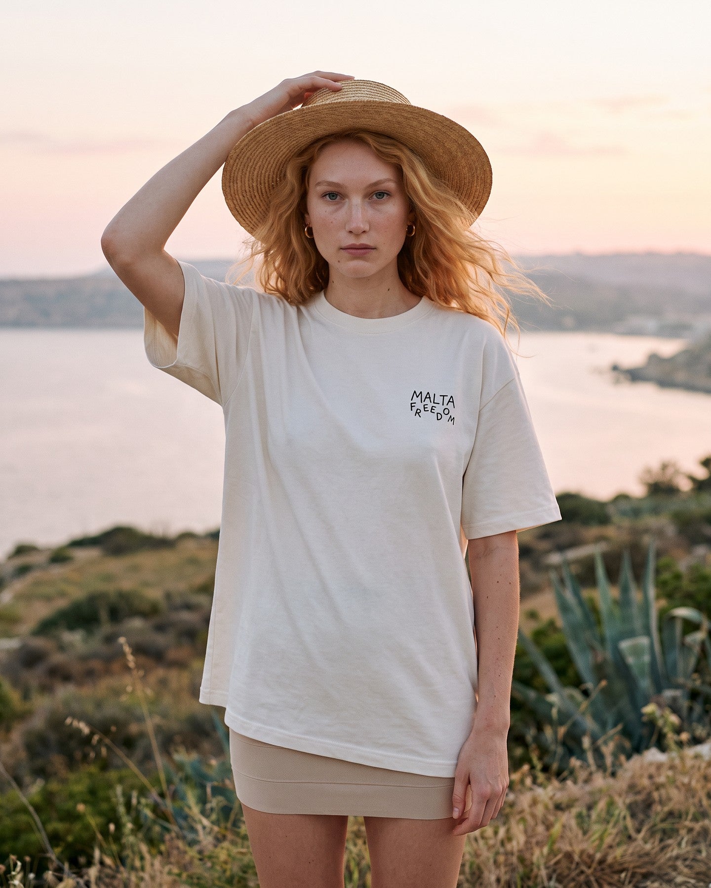 Mulher usando a camiseta oversized MALTA® flying off-white numa encosta sobre a baía ao pôr do sol