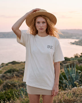 Mulher usando a camiseta oversized MALTA® flying off-white numa encosta sobre a baía ao pôr do sol