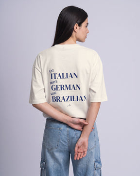 Camiseta oversized feminina MALTA® kiss brazilian off-white vista de costas no estúdio, com a frase “eat italian, drive german, kiss brazilian”