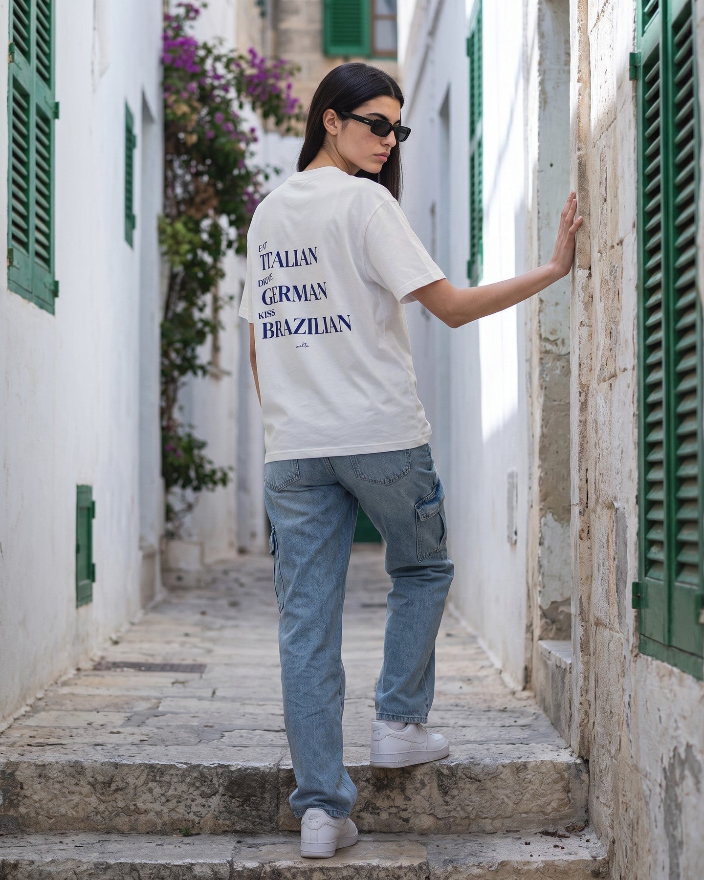 Mulher de costas usando a camiseta oversized MALTA® kiss brazilian off-white numa ruela caiada