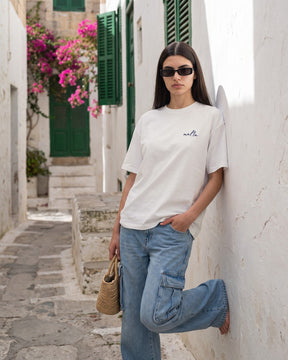 Mulher usando a camiseta oversized MALTA® kiss brazilian off-white numa ruela caiada