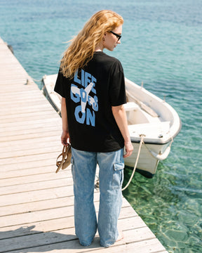 Mulher de costas usando a camiseta oversized MALTA® life goes on preta num píer sobre água turquesa