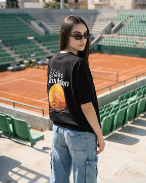 Mulher de costas usando a camiseta oversized MALTA® life is a simulation preta na arquibancada de um clube de tênis