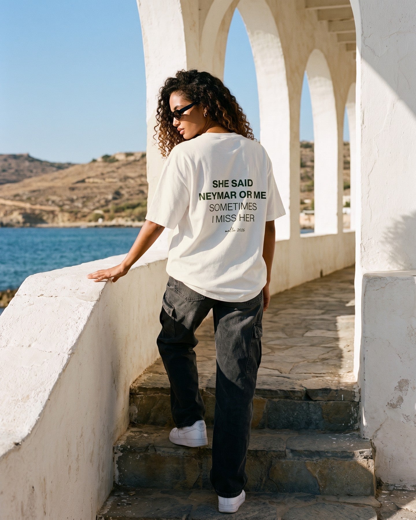 Mulher de costas usando a camiseta oversized MALTA® neymar or me off-white numa galeria de arcos caiados