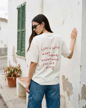 Mulher de costas usando a camiseta oversized MALTA® nobody knows off-white numa parede caiada com janela verde