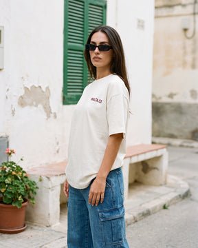 Mulher usando a camiseta oversized MALTA® nobody knows off-white numa parede caiada com janela verde