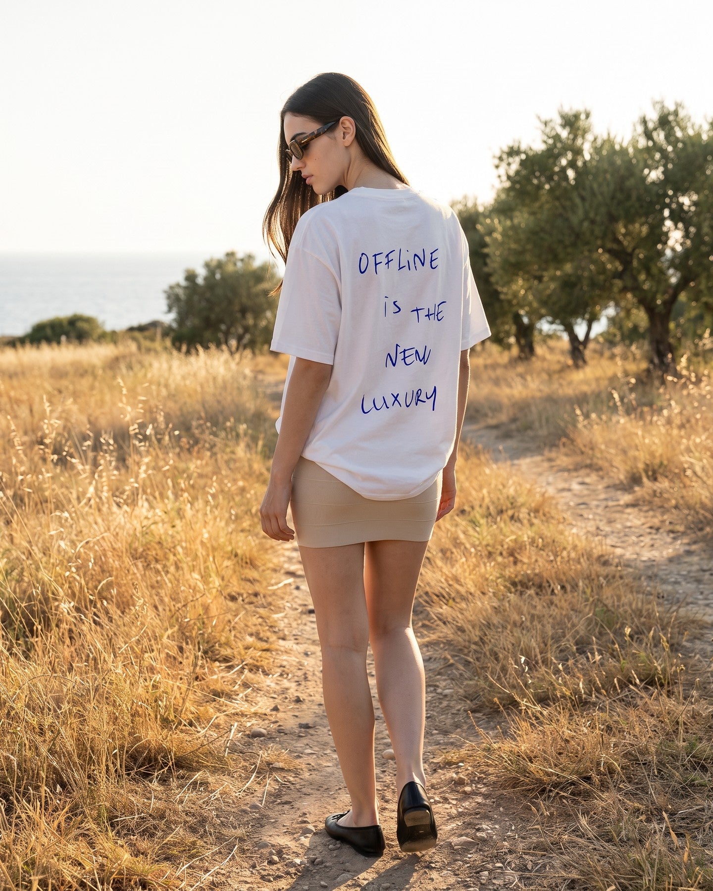 Mulher de costas usando a camiseta oversized MALTA® offline luxury off-white num campo com oliveiras