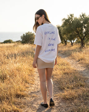 Mulher de costas usando a camiseta oversized MALTA® offline luxury off-white num campo com oliveiras