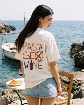 Mulher de costas usando a camiseta oversized MALTA® pasta sex wine off-white numa mesa posta à beira-mar