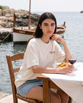 Mulher usando a camiseta oversized MALTA® pasta sex wine off-white numa mesa posta à beira-mar