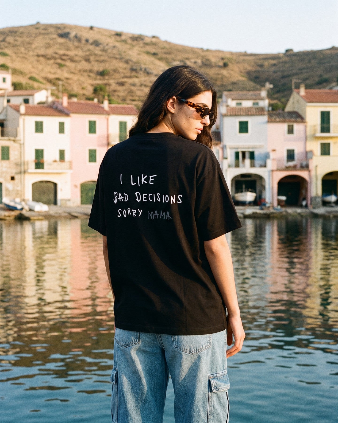 Mulher de costas usando a camiseta oversized MALTA® bad decisions preta numa vila de pescadores