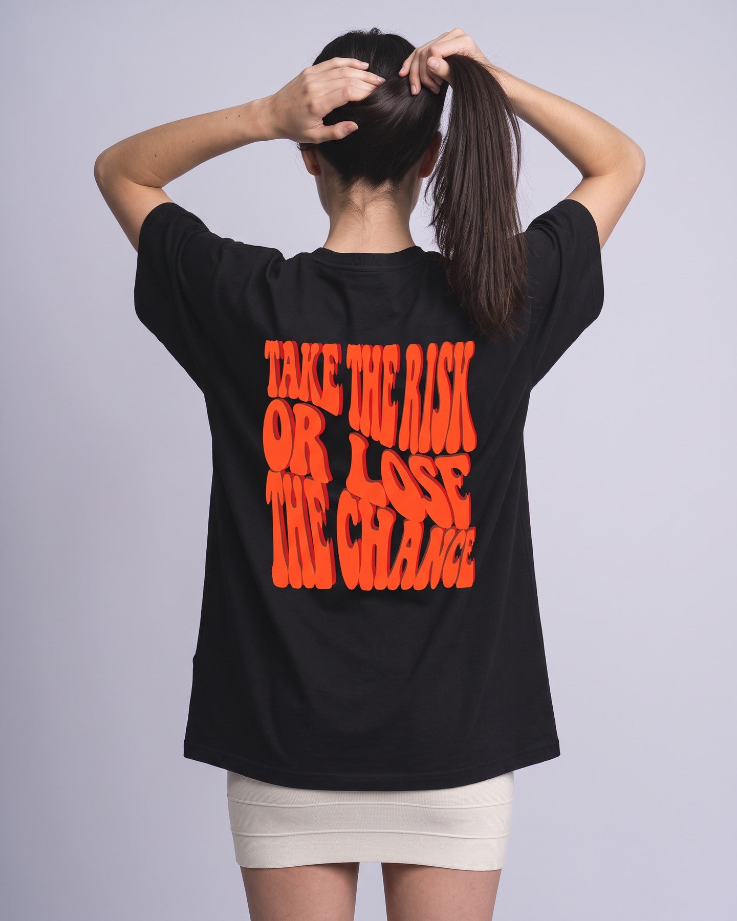 Camiseta oversized feminina MALTA® take the risk preta vista de costas no estúdio, com a frase “take the risk or lose the chance”