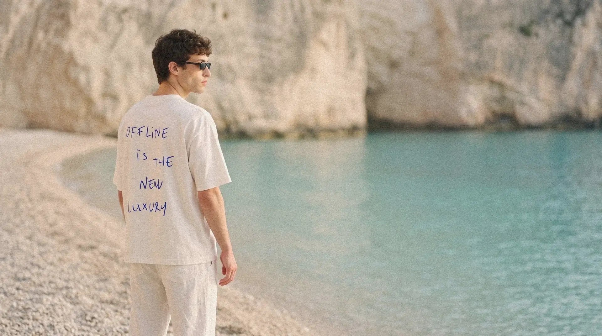 Homem de costas numa enseada de pedras claras e água turquesa, vestindo a camiseta MALTA offline is the new luxury