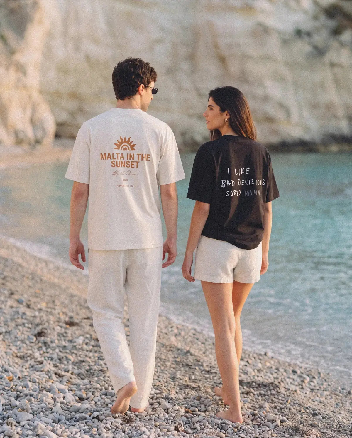 Casal de costas caminhando numa praia de pedras claras, com as camisetas MALTA malta in the sunset e i like bad decisions, sorry mama